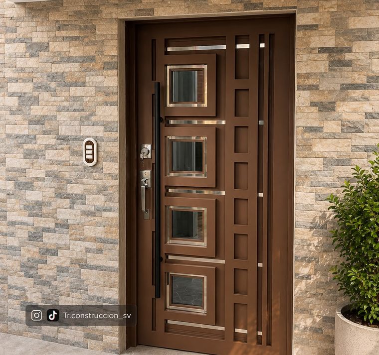 Puerta metálica color bronce con cuatro vidrios cuadrados enmarcados en acero inoxidable y panel lateral calado, sobre fachada de piedra laja